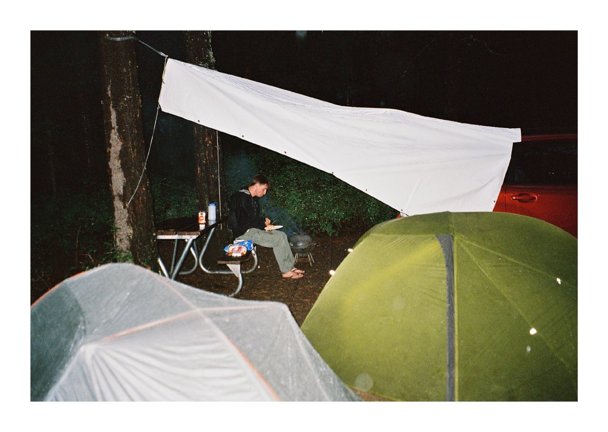 A camper eating beneath a tarp among tents at a rainy forest campsite