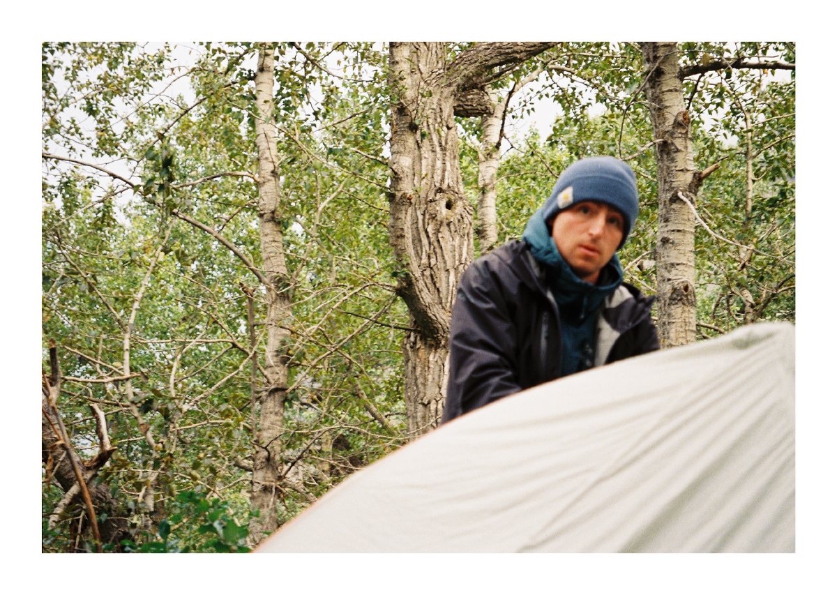 A camper in a blue beanie pitching a tent among leafy trees