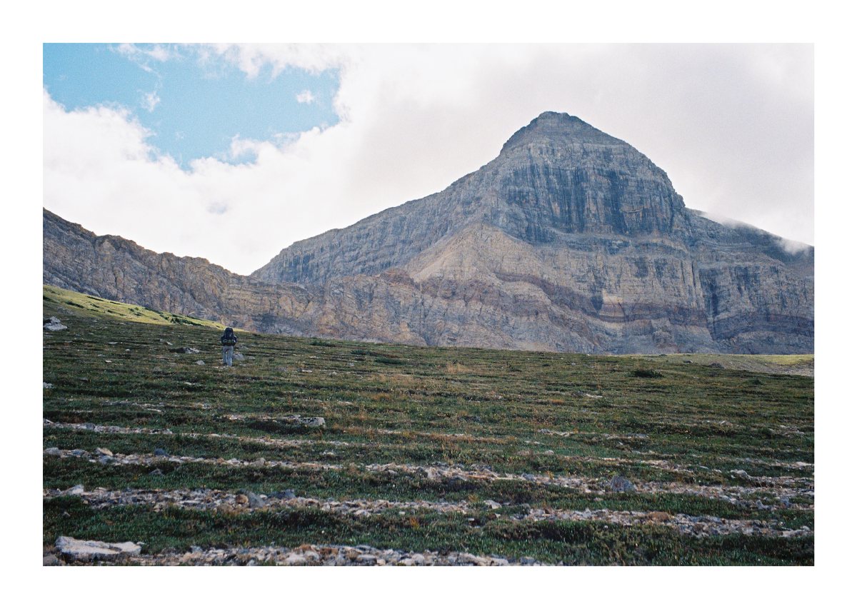 A backpacker crossing an alpine meadow below a massive layered peak