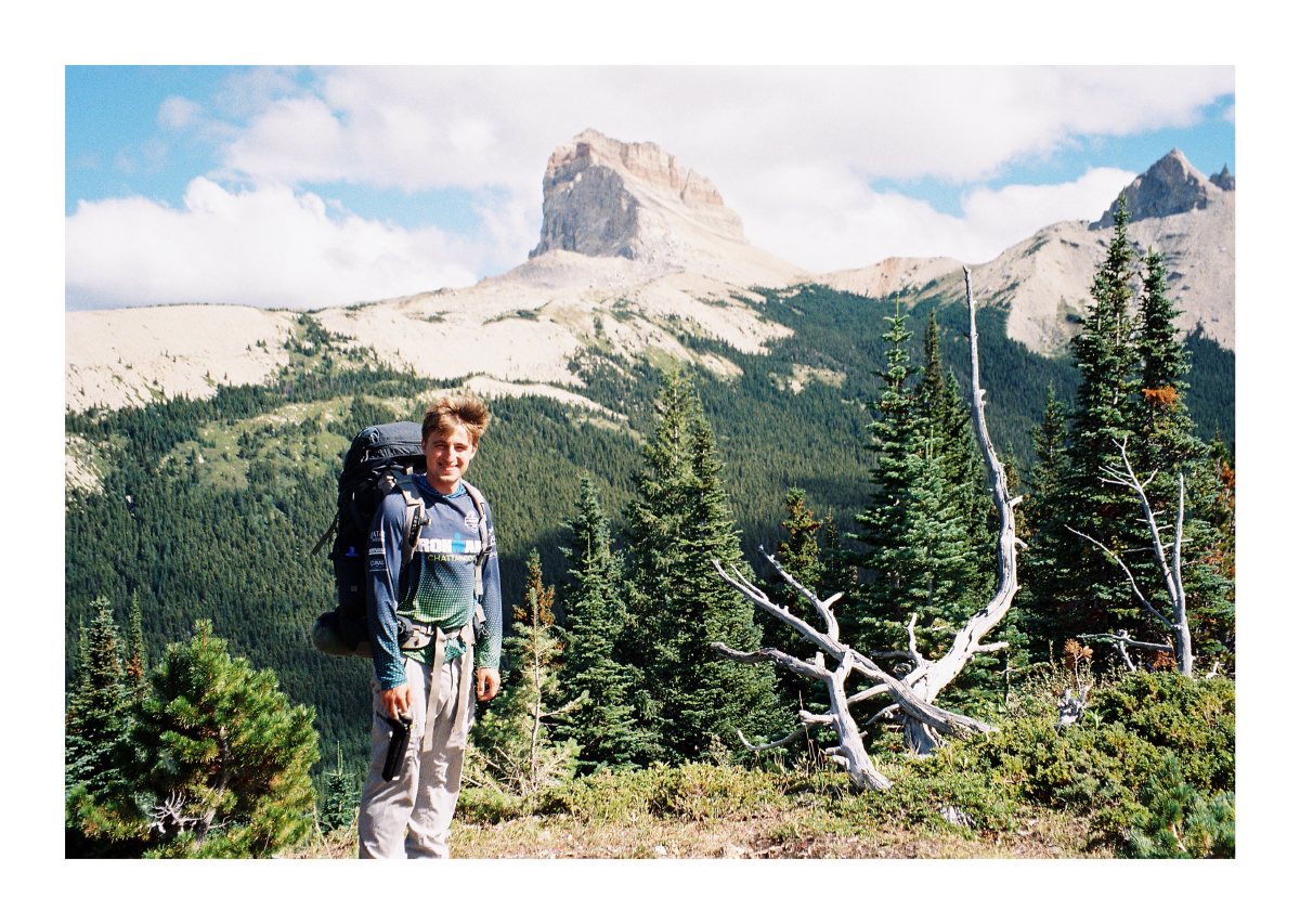 A smiling backpacker posing before a rugged mountain and evergreen valley