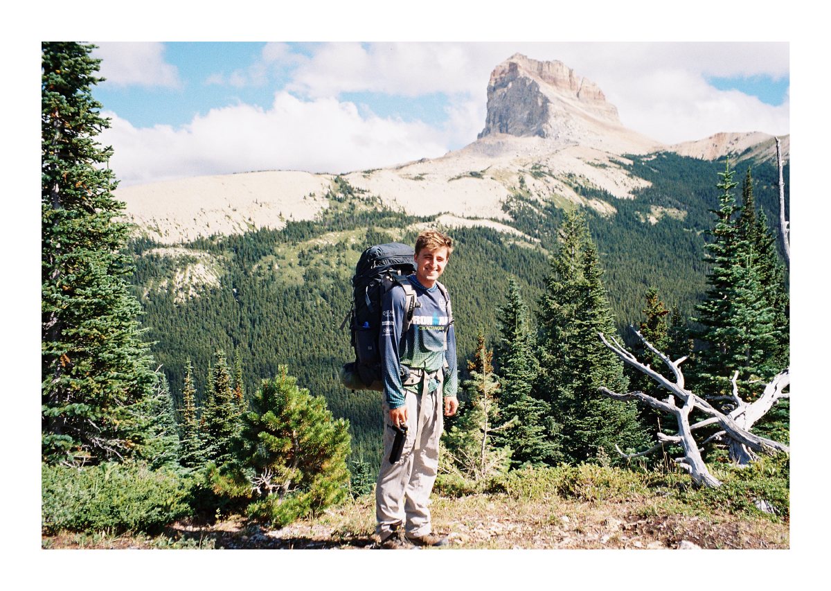 A backpacker posing among evergreens with a pale rock peak behind him
