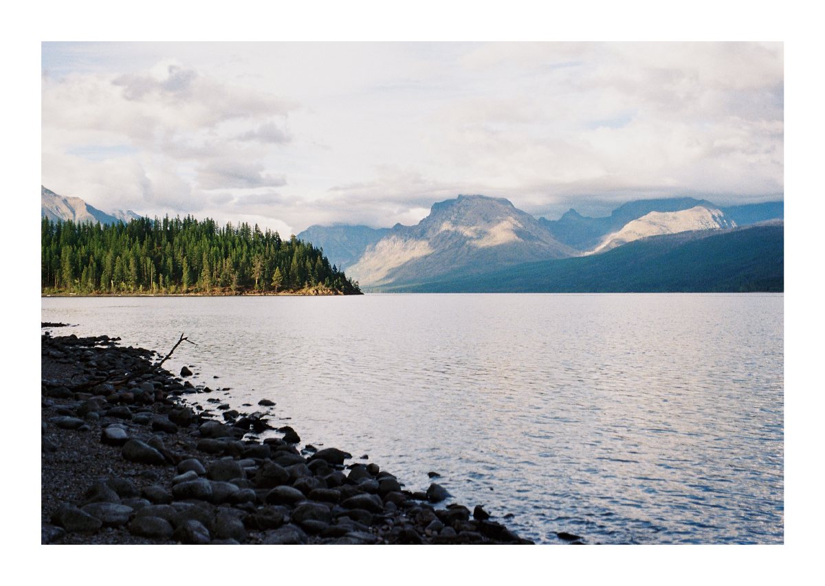 A calm mountain lake bordered by a rocky shore, evergreen peninsula, and distant peaks